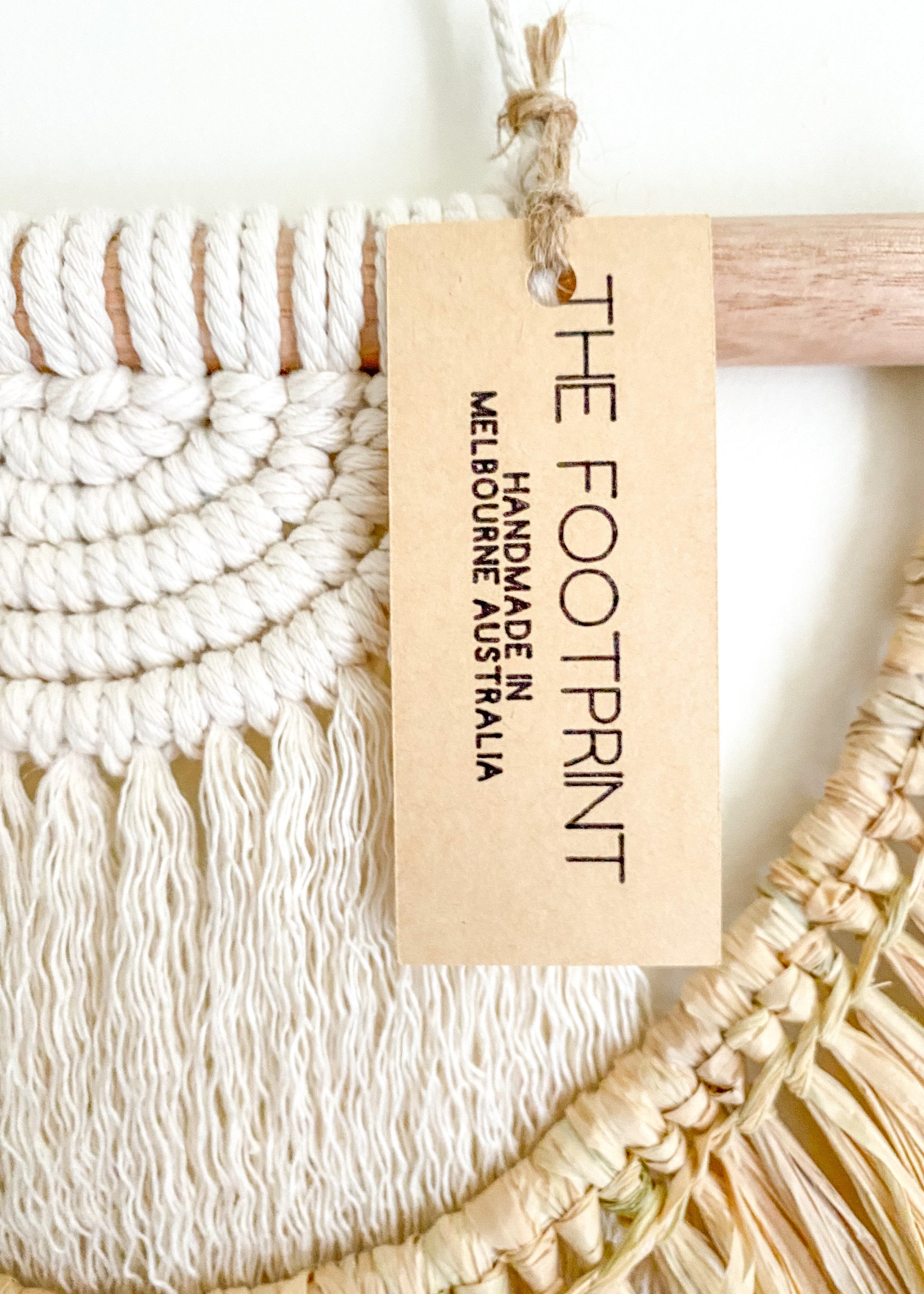 Close up view of the product tag of macrame and raffia wall hanging. Product tag reads - The Footprint: Handmade in Melbourne Australia. 