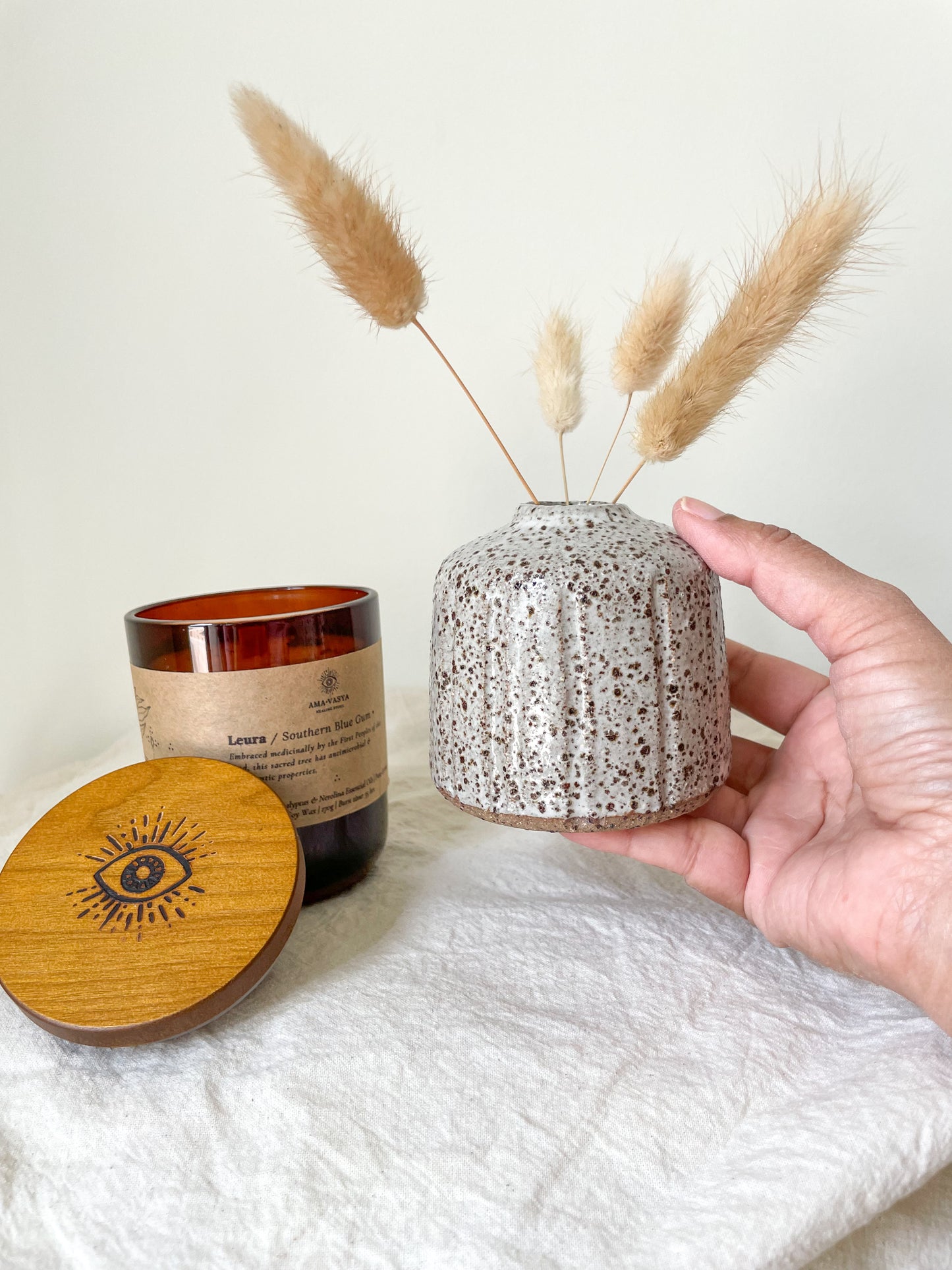 Leura soywax candle and Hazel small handmade ceramic vase with dried bunny tails in it. Small ceramic vase is held in a hand