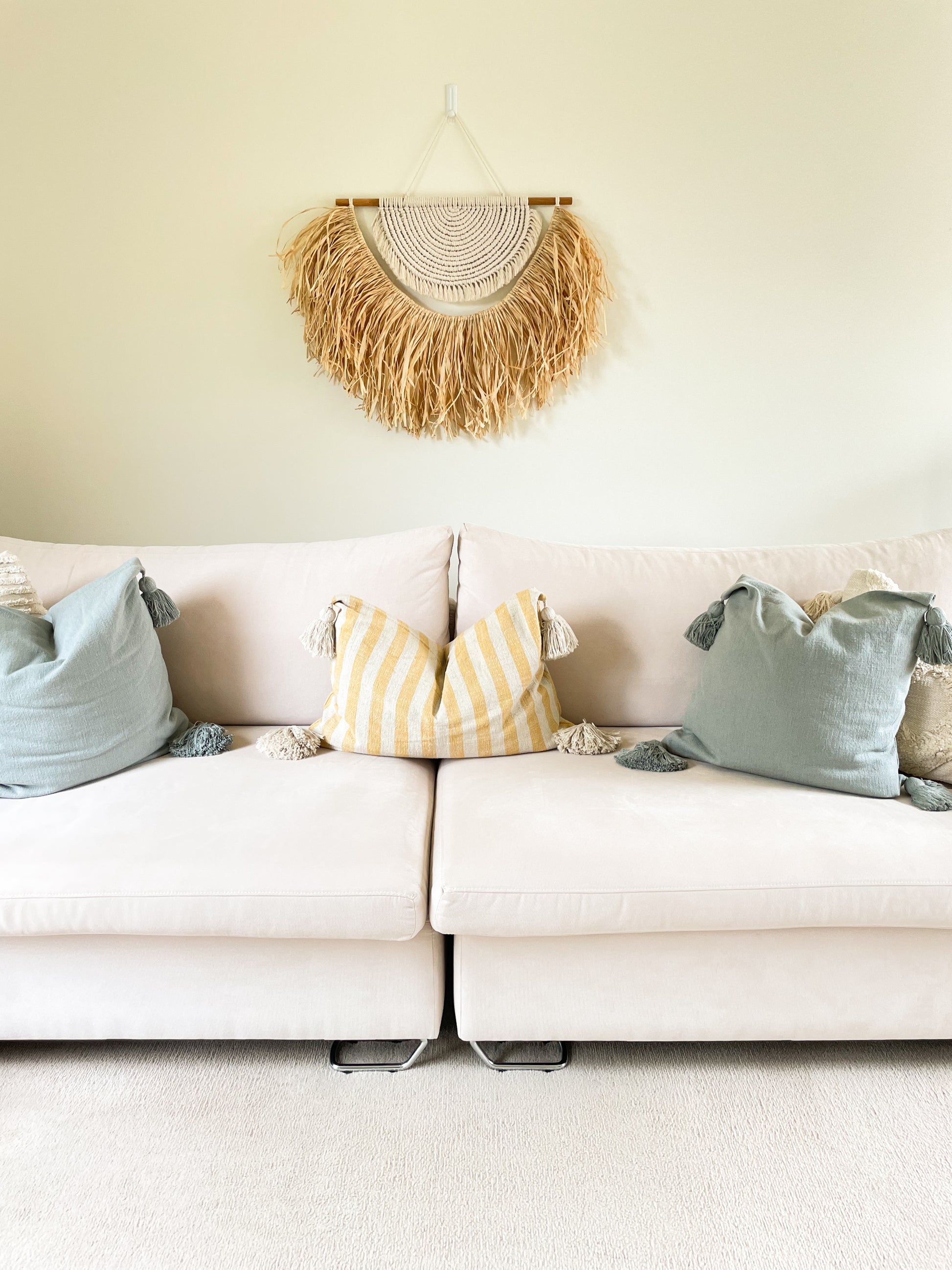 Large macrame and raffia wall hanging hanged on a wall above a large sofa styled with coastal style cushions