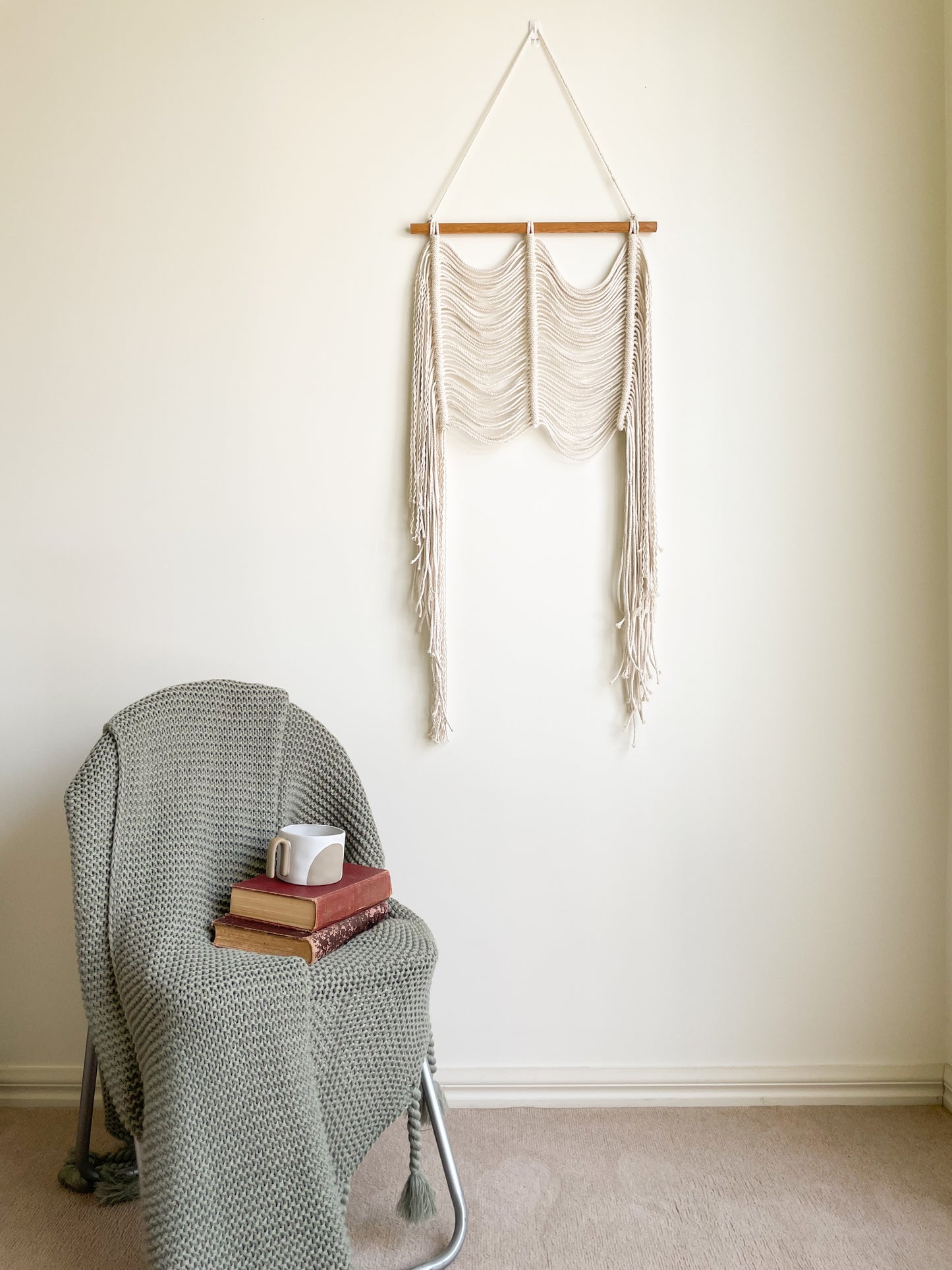 Modern macrame wall hanging hanged on a wall above a chair styled with a throw and vintage books