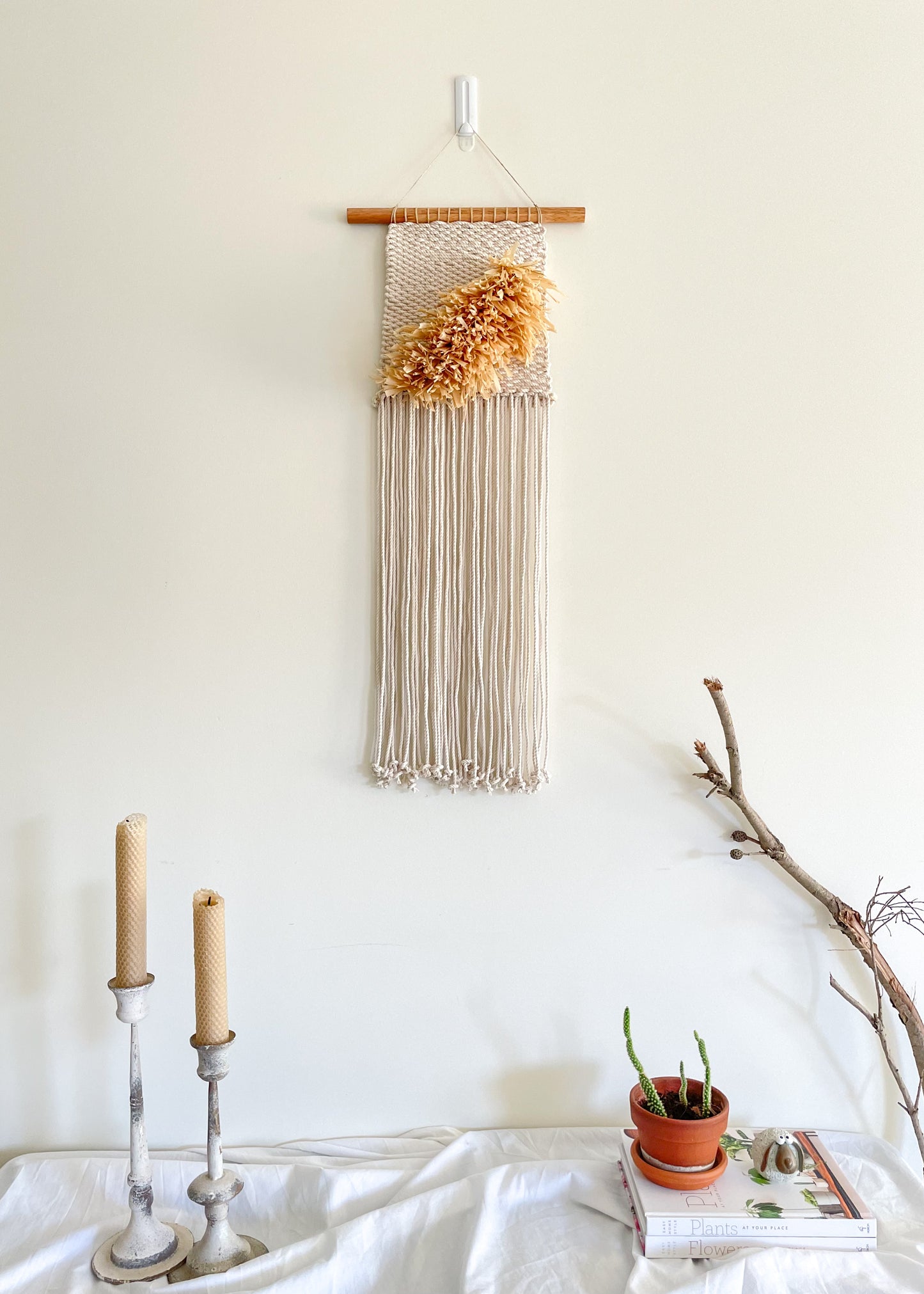 Raffia and cotton handwoven wall hanging hanged on a wall above a sideboard. styled with candles, books and a cactus potted plant
