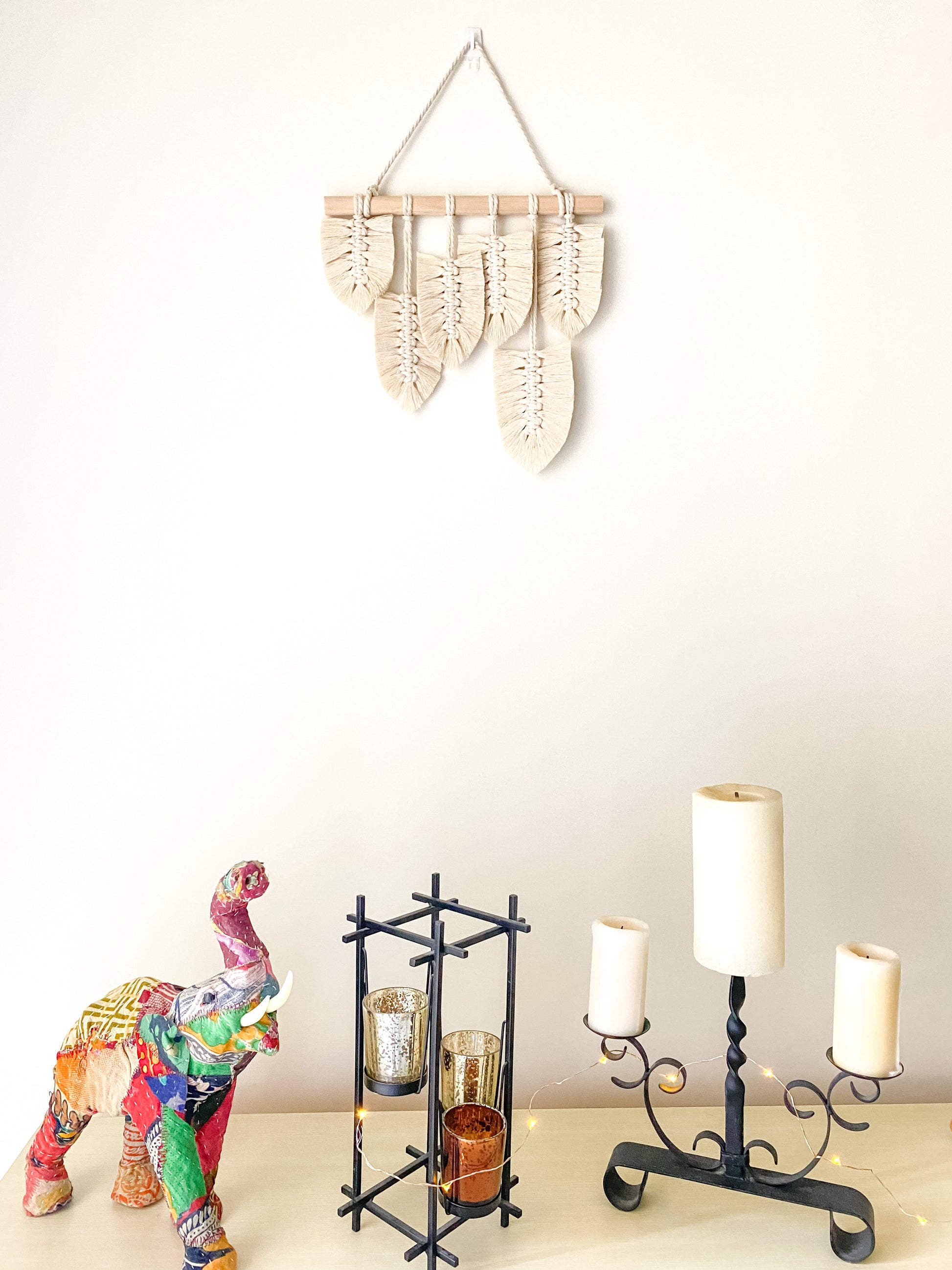 Natural white feather macrame wall hanging hanged on a wall above a side board buffet decorated with two candle holders, fabric elephant and fairy lights. 