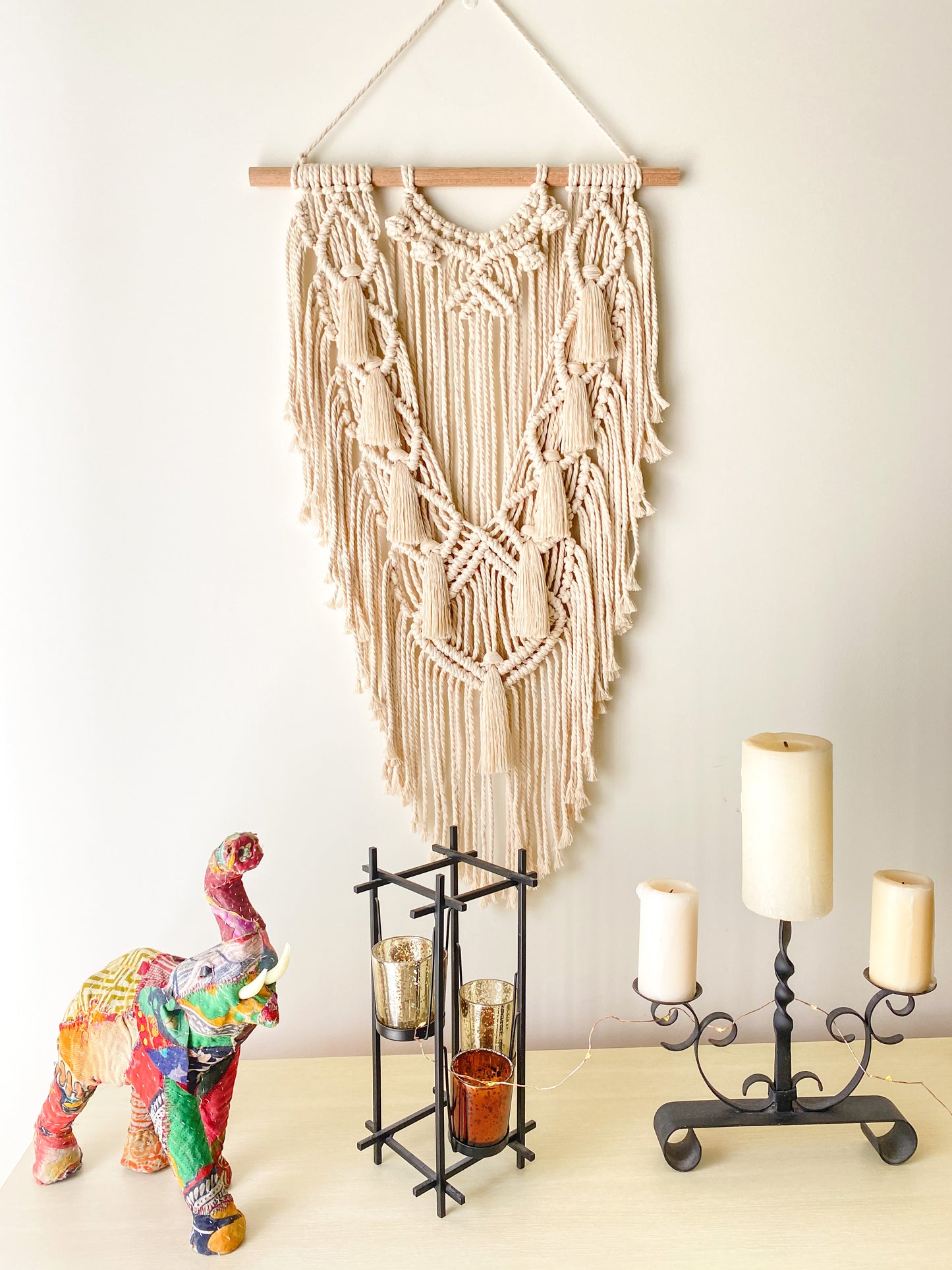 Natural white macrame wall hanging with tassels hanged on a wall above a table styled with two candles holders and a fabric elephant