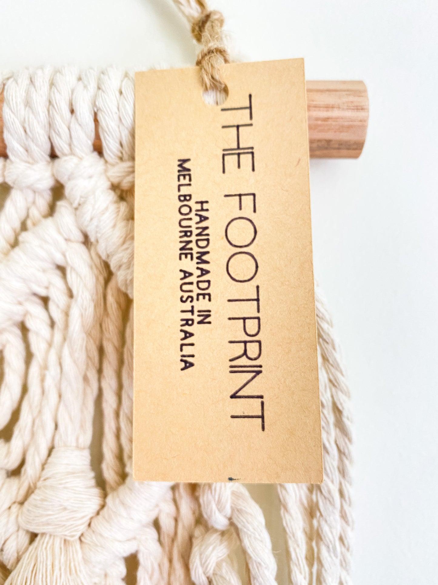 Closeup photo of a natural off white macrame wall hanging with the product tag on. Product tag reads The Footprint handmade in Melbourne, Australia