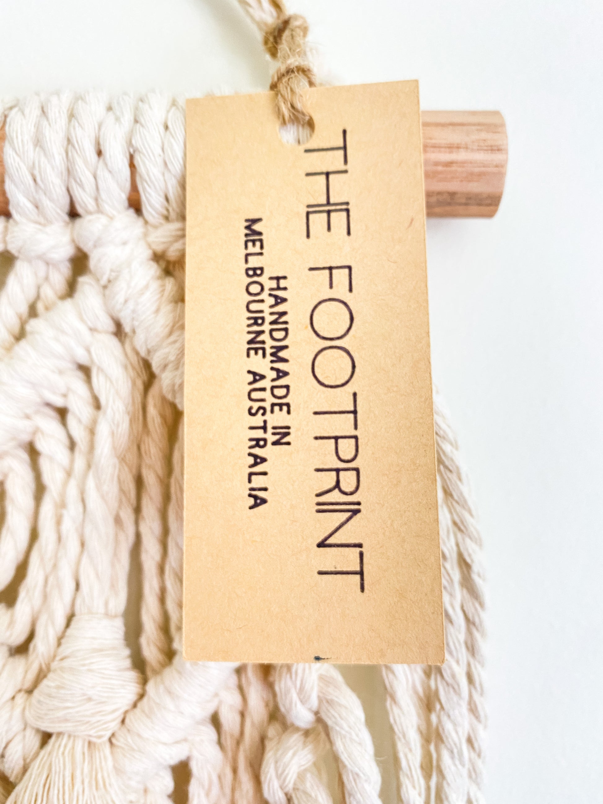 Closeup photo of a natural off white macrame wall hanging with the product tag on. Product tag reads The Footprint handmade in Melbourne, Australia
