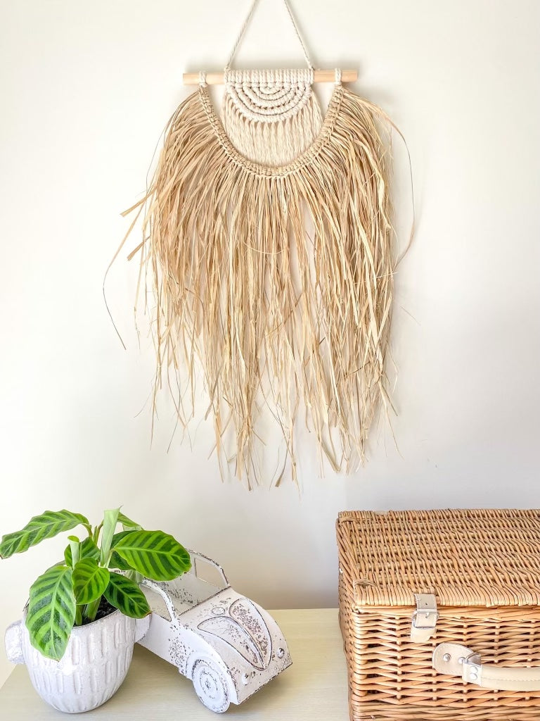 Macrame and raffia wall hanging hanged on the wall above a buffet styled with a rattan picnic basket, indoor plant and a rustic car ornament