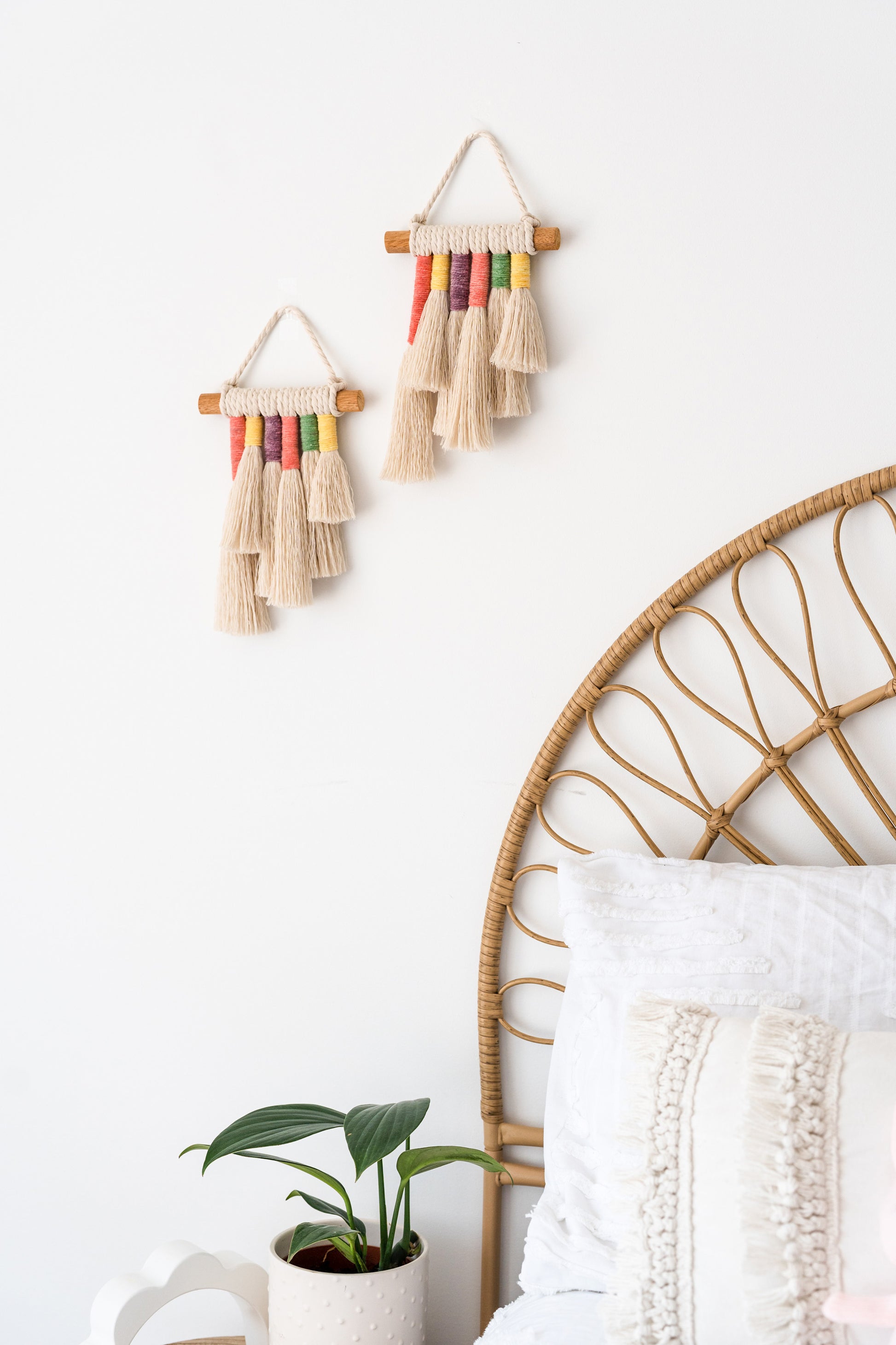 Two small macrame rainbow wall hangings hanged on a white wall of a kids bedroom. Above the bed. Bedside table next to the table with a white decor piece and a potted plant