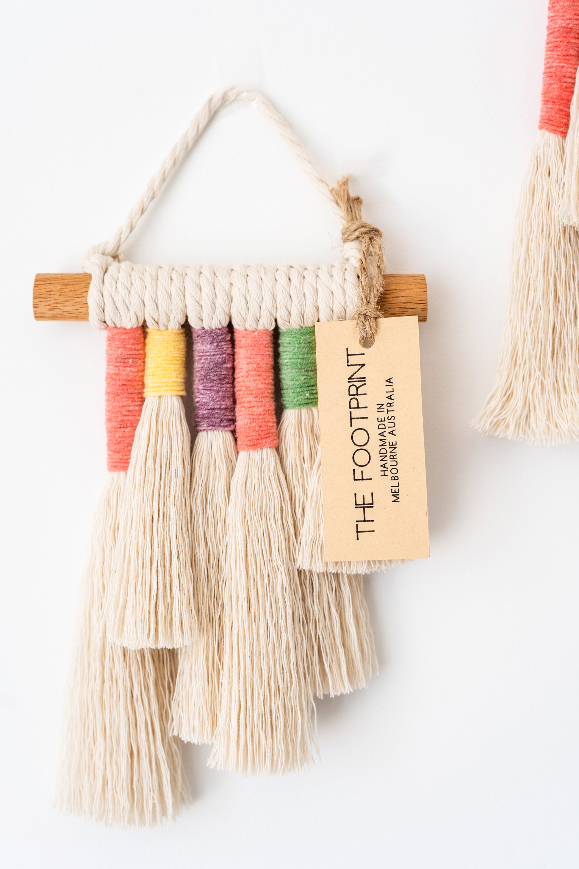 Close up photo of a mini rainbow macrame wall hanging with a product tag on it. Product tag reads The Footprint handmade in Melbourne, Australia