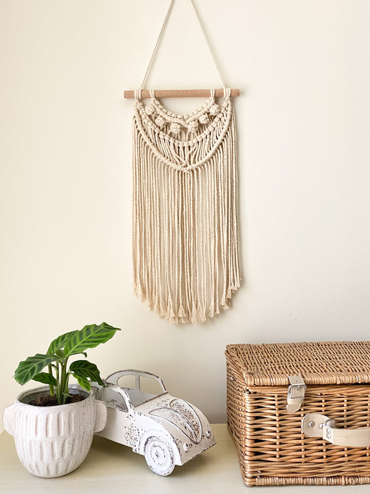 Natural off white macrame tassels hanging hanged on the wall above a table styled with an indoor plant white coastal themed toy car, rattan picnic basket.