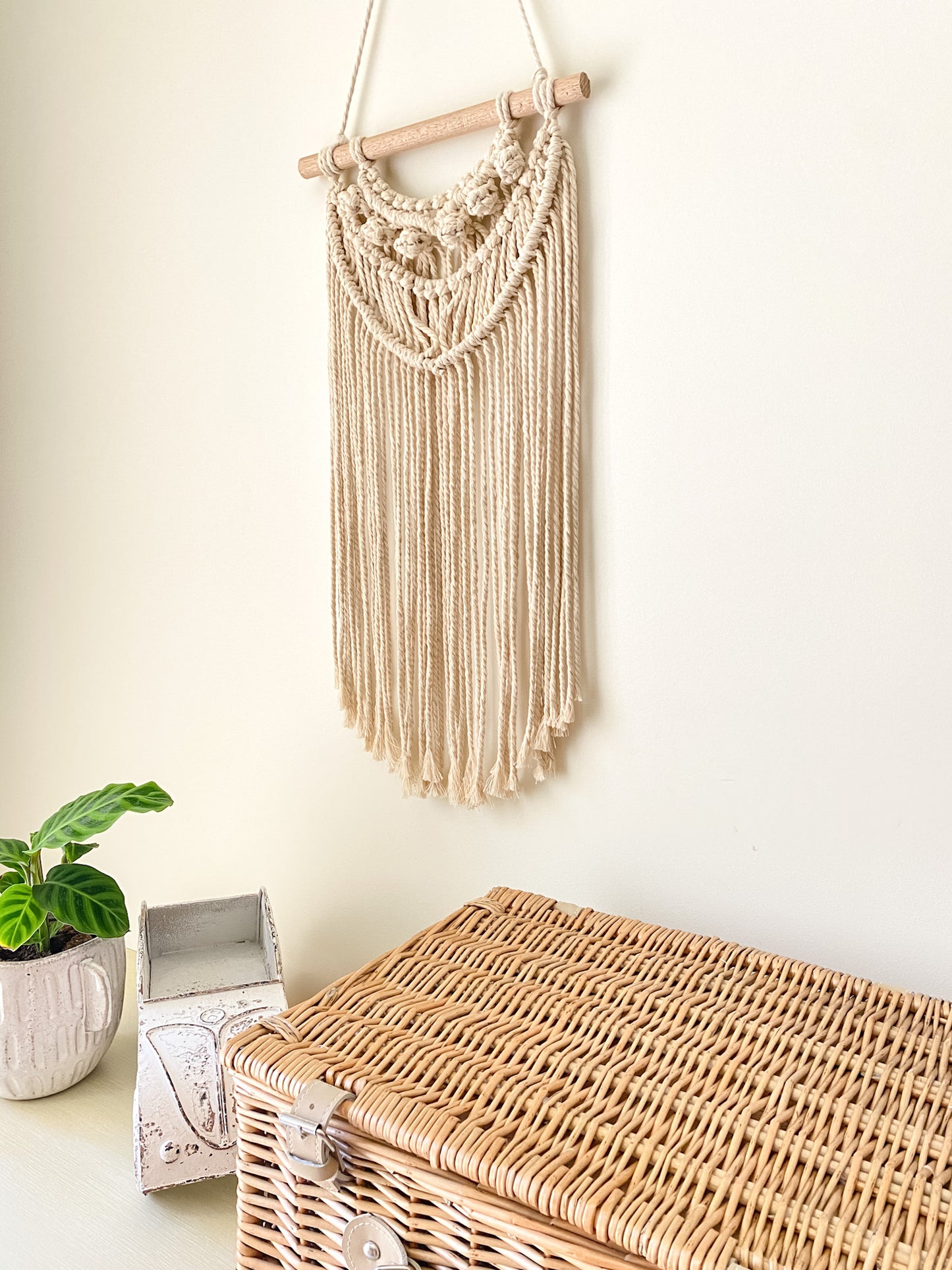 Side view of a natural off white macrame tassels hanging hanged on the wall above a table styled with an indoor plant white coastal themed toy car, rattan picnic basket.